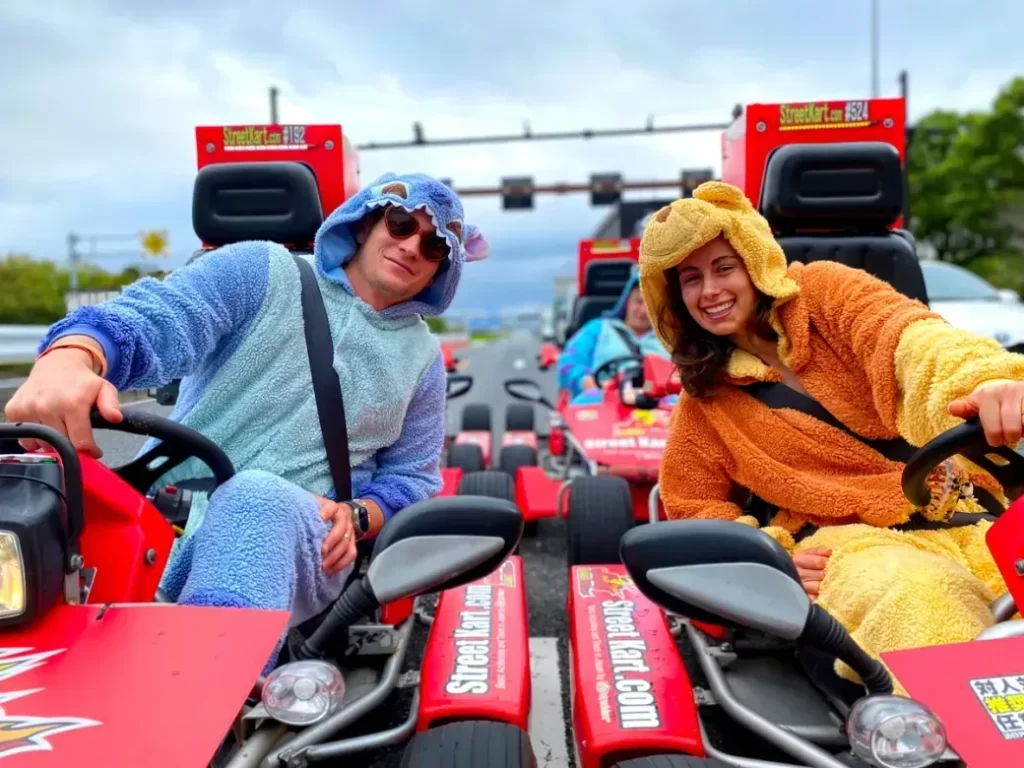 IMG_6138 - StreetKart Two people wearing colorful animal onesies pose on red go-karts, smiling at the camera on a race track.