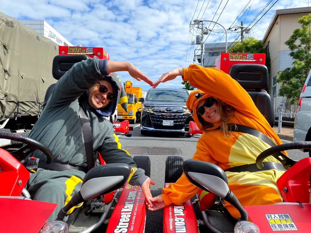 Two people in go-karts on a street form a heart with their arms, smiling in costumes (gray suit and orange onesie).